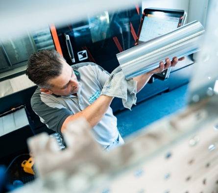 Ein Arbeiter hält ein großes Metallrohr in der Hand und steht in einer Werkstatt oder Fabrik.