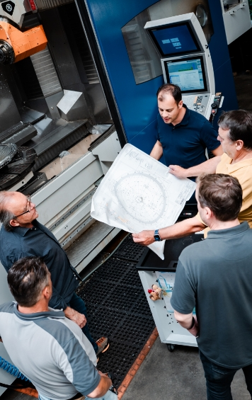 Fünf Personen besprechen technische Pläne vor einer CNC-Maschine in einer industriellen Umgebung.