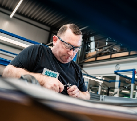 Ein Mitarbeiter in einer Werkstatt arbeitet konzentriert mit Werkzeug, trägt Schutzbrille und ein schwarzes T-Shirt.