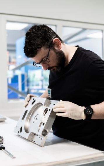 Ein Mann mit Brille und Handschuhen untersucht ein Metallteil in einer Werkstatt.