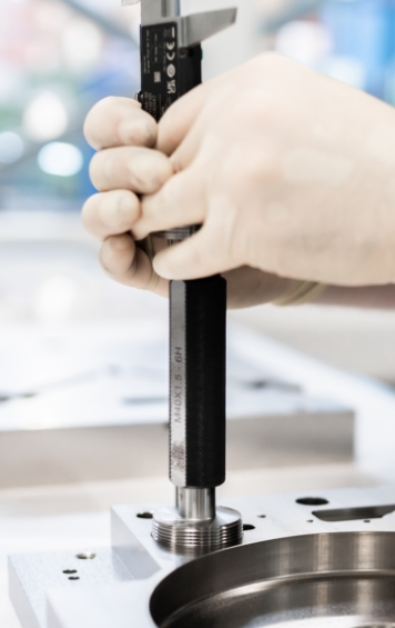 Eine Hand mit Handschuhen hält einen Messschieber, während sie eine Metallkomponente auf einem Tisch misst.