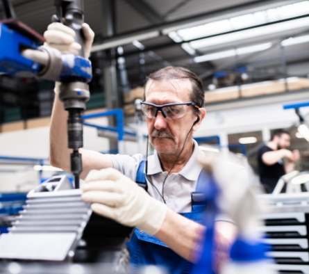 Ein älterer Mann in Schutzbrille und Handschuhen arbeitet an einer Maschine in einer Fabrik.