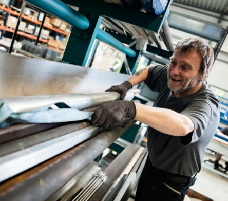 Ein Arbeiter lächelt, während er Metallrohre in einer Werkstatt bearbeitet.