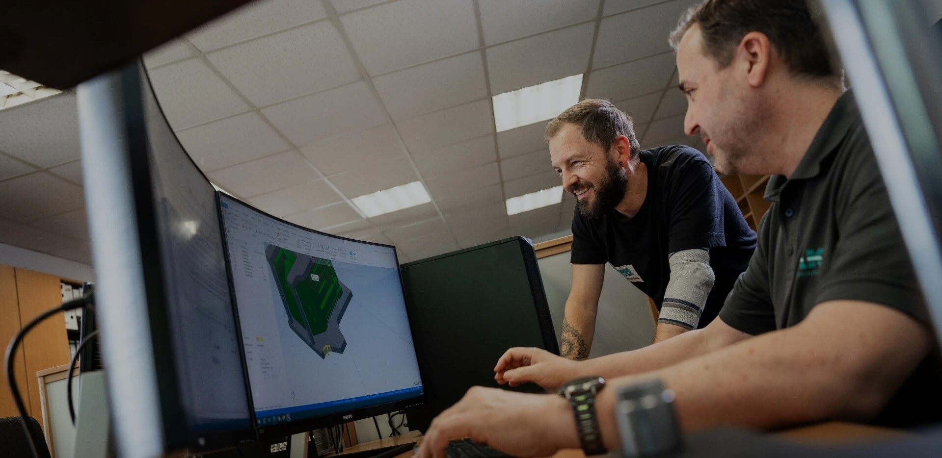 Zwei Männer arbeiten an einem Computer, der ein 3D-Modell zeigt. Sie lachen und scheinen kollaborativ zu sein.