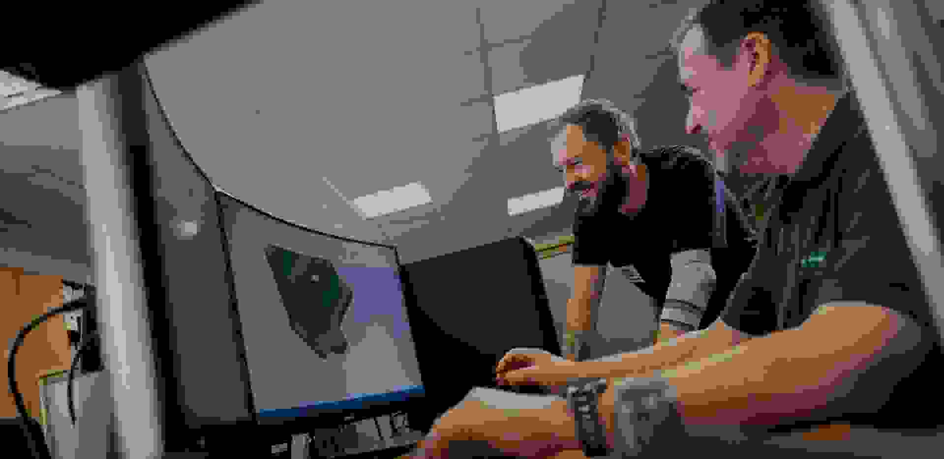 Zwei Männer arbeiten an einem Computer, der ein 3D-Modell zeigt. Sie lachen und scheinen kollaborativ zu sein.