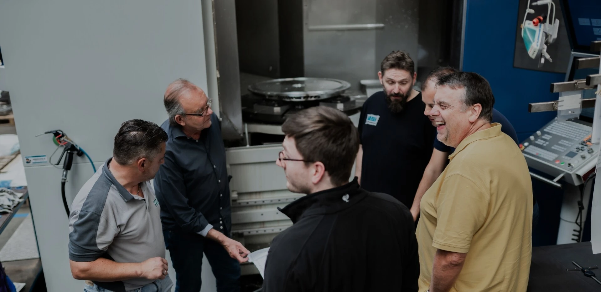 Eine Gruppe von sechs Männern steht um eine Maschine und diskutiert. Sie schauen interessiert auf ein Dokument und scheinen etwas zu erklären oder zu lernen.