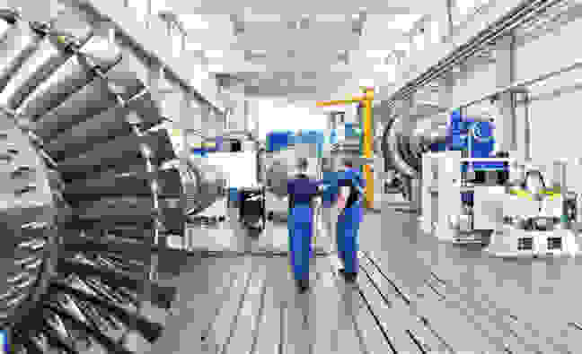 Zwei Arbeiter gehen in einer modernen Fertigungshalle zwischen großen Turbinen.