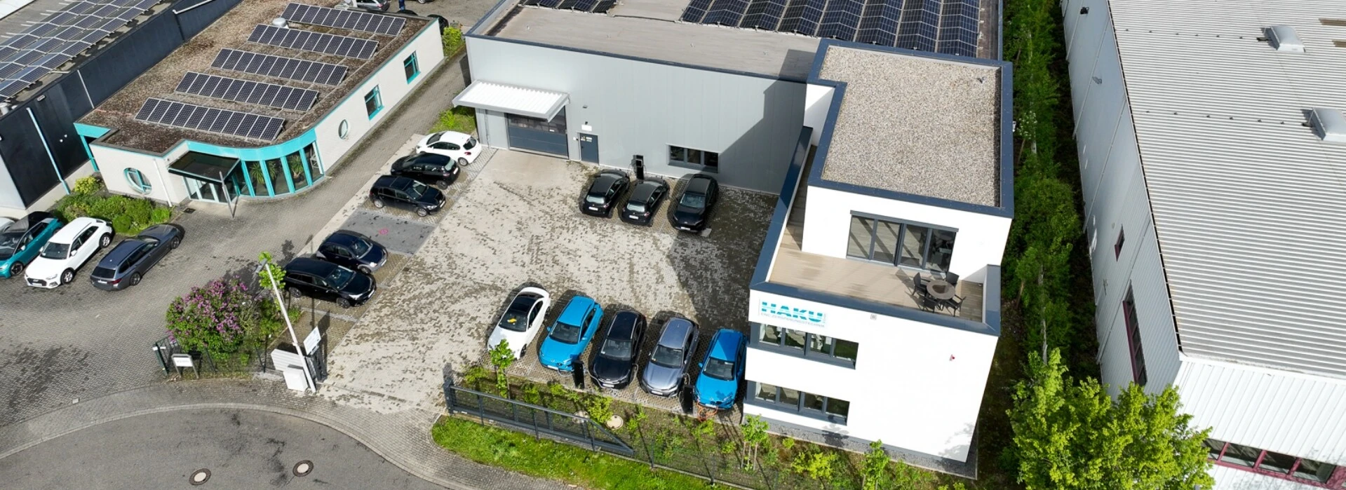 Eine Luftaufnahme eines modernen Gebäudes mit Solaranlagen auf dem Dach, umgeben von einem Parkplatz mit mehreren Autos und Pflanzen.
