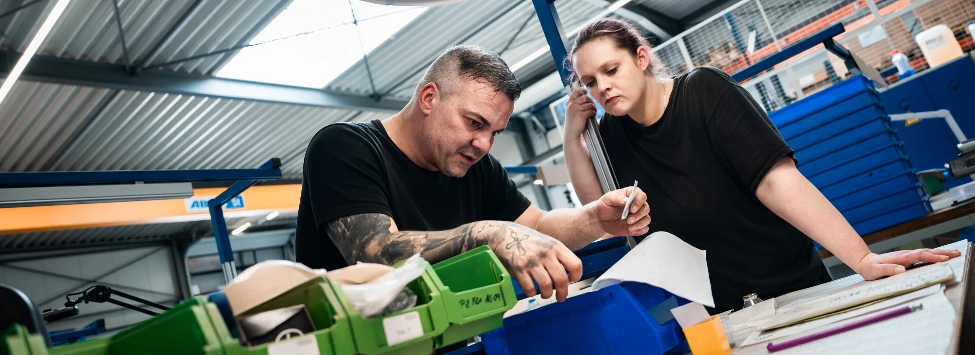 Zwei Personen arbeiten in einer Industrieumgebung, Blick auf eine Arbeitsfläche mit Werkzeugen und Materialien.