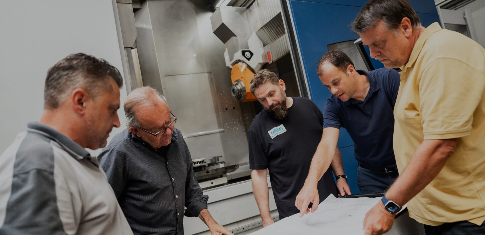 Fünf Männer betrachten gemeinsam technische Pläne vor einer Maschinenpresse in einer industriellen Umgebung.