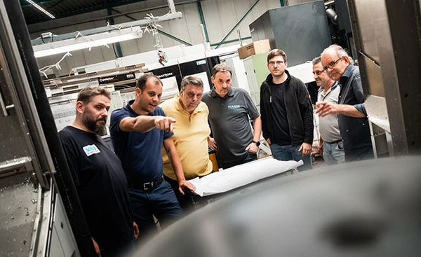 Gruppe von Männern in einer Werkstatt, die an einer Maschine stehen und diskutieren. Eine große Metallplatte ist im Vordergrund sichtbar.