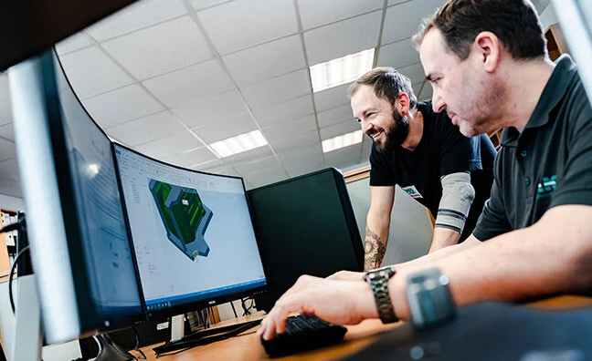 Drei Männer sitzen an einem Tisch, schauen auf Unterlagen und diskutieren. Ein Mann hält ein Blatt Papier, während ein anderer Notizen macht.