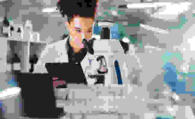 Eine Wissenschaftlerin im Labor arbeitet mit einem Mikroskop und einem Tablet. Im Hintergrund sind Reagenzgläser und Labormaterialien zu sehen.