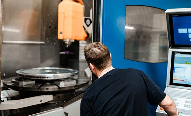 Eine Maschinenbearbeitung mit einem Werkzeug, das einen zylindrischen Metallteil fräst. Helles Licht und reflektierende Oberflächen sind sichtbar.