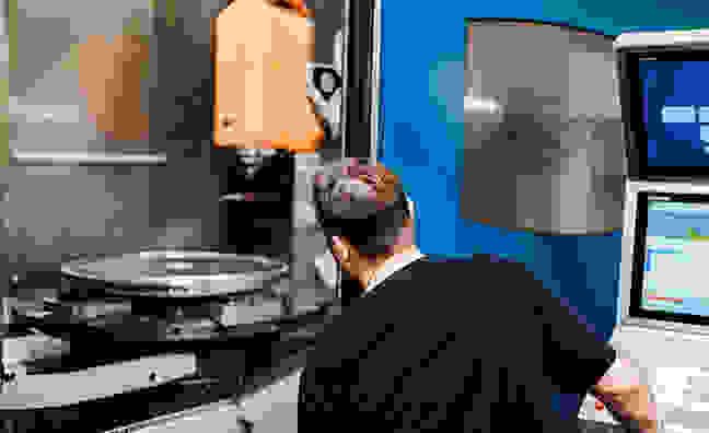 Eine Maschinenbearbeitung mit einem Werkzeug, das einen zylindrischen Metallteil fräst. Helles Licht und reflektierende Oberflächen sind sichtbar.