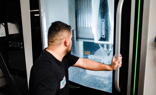 Ein Arbeiter in blauer Latzhose sitzt an einer Maschine und montiert Teile, während er einen Roboterarm beobachtet.