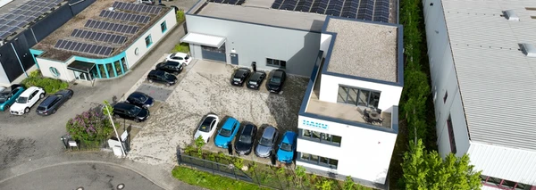 Eine Luftaufnahme eines modernen Gebäudes mit Solaranlagen auf dem Dach, umgeben von einem Parkplatz mit mehreren Autos und Pflanzen.