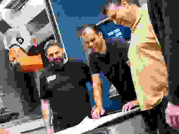 Eine Gruppe von fünf Personen steht um einen Tisch und betrachtet einen Plan. Im Hintergrund ist eine Maschinenwerkstatt mit Maschinen sichtbar.