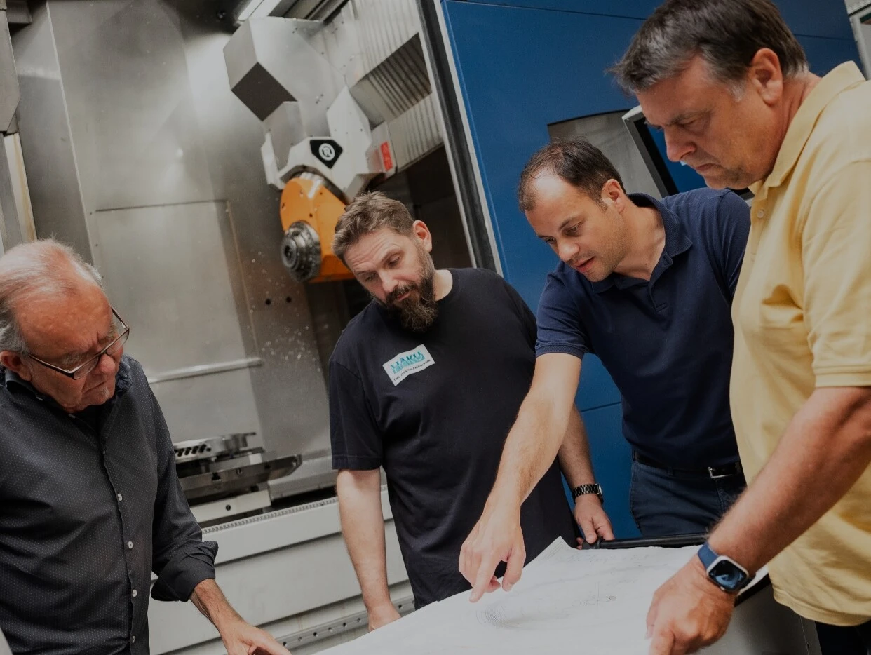 Fünf Männer betrachten gemeinsam technische Pläne vor einer Maschinenpresse in einer industriellen Umgebung.