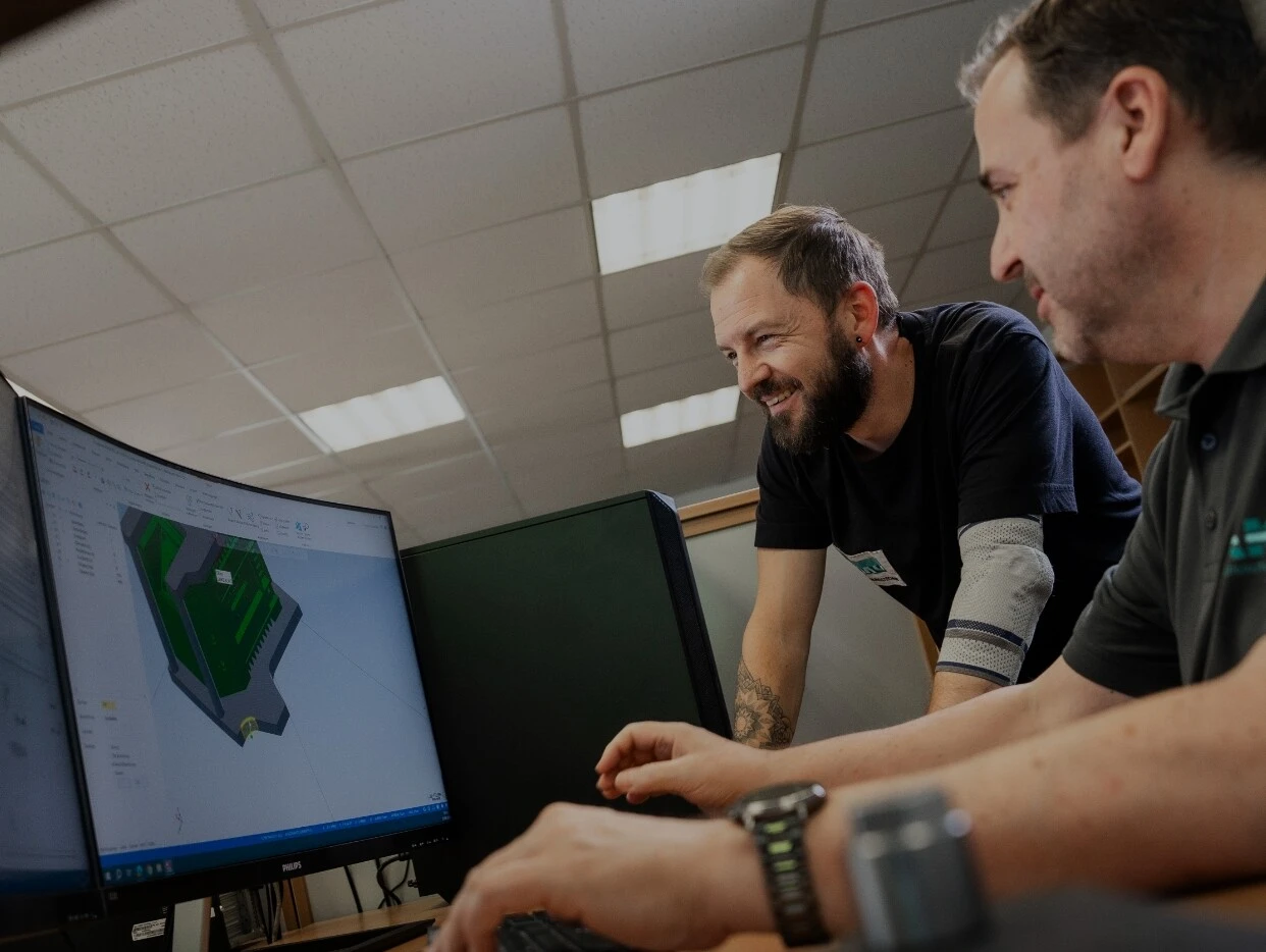 Zwei Männer arbeiten an einem Computer, der ein 3D-Modell zeigt. Sie lachen und scheinen kollaborativ zu sein.