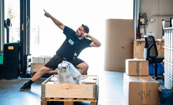 Ein Mann in einem Lager posiert dynamisch neben einer Holzkiste und Kartons.