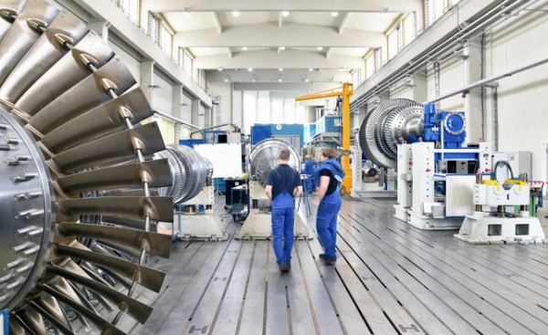 Zwei Arbeiter gehen in einer modernen Fertigungshalle zwischen großen Turbinen.
