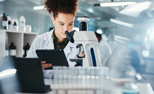 Eine Wissenschaftlerin im Labor arbeitet mit einem Mikroskop und einem Tablet. Im Hintergrund sind Reagenzgläser und Labormaterialien zu sehen.