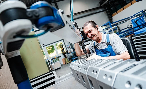 Ein Mann arbeitet an einer Maschine und hält ein Werkzeug in der Hand. Im Hintergrund sind Bedienelemente sichtbar.